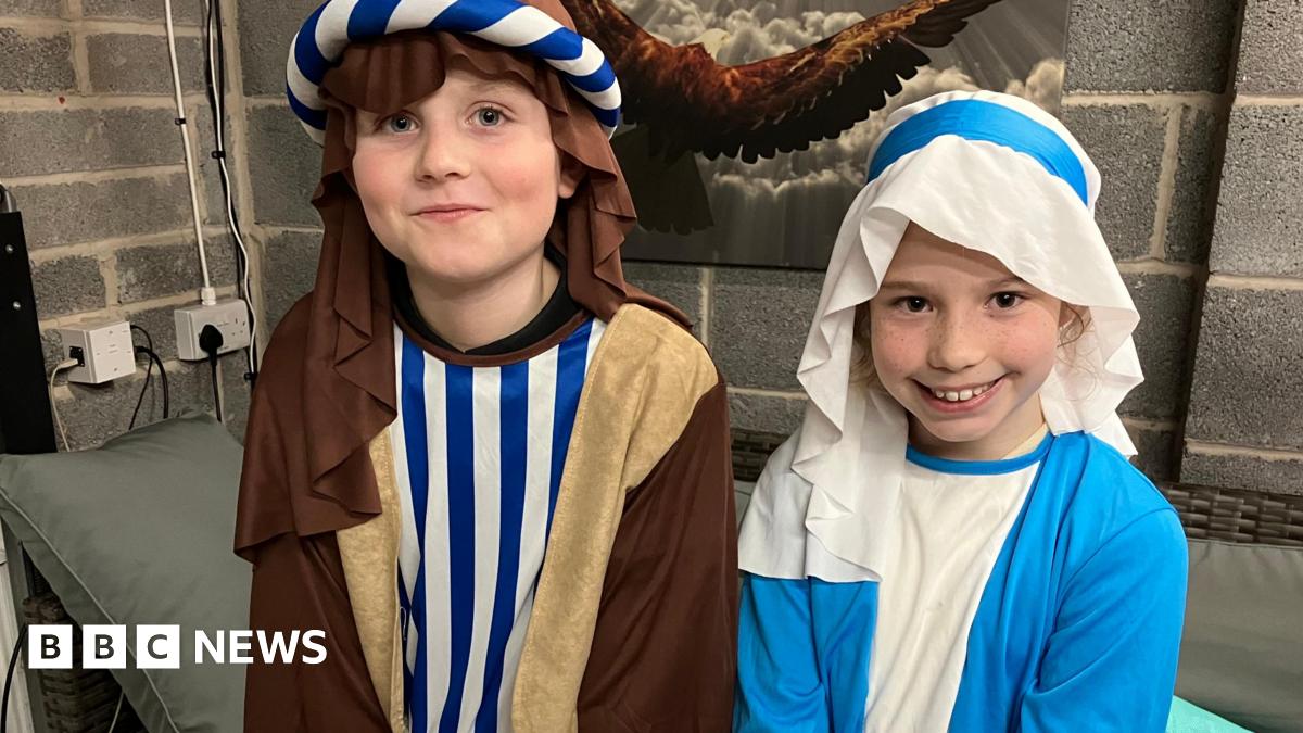 Oksi, who plays Joseph, and Elsie, who is Mary, are standing next to each other wearing traditional nativity costumes for the production. They are smiling at the camera. There is a cushion behind them and they are standing in a garage which has a picture on the wall.