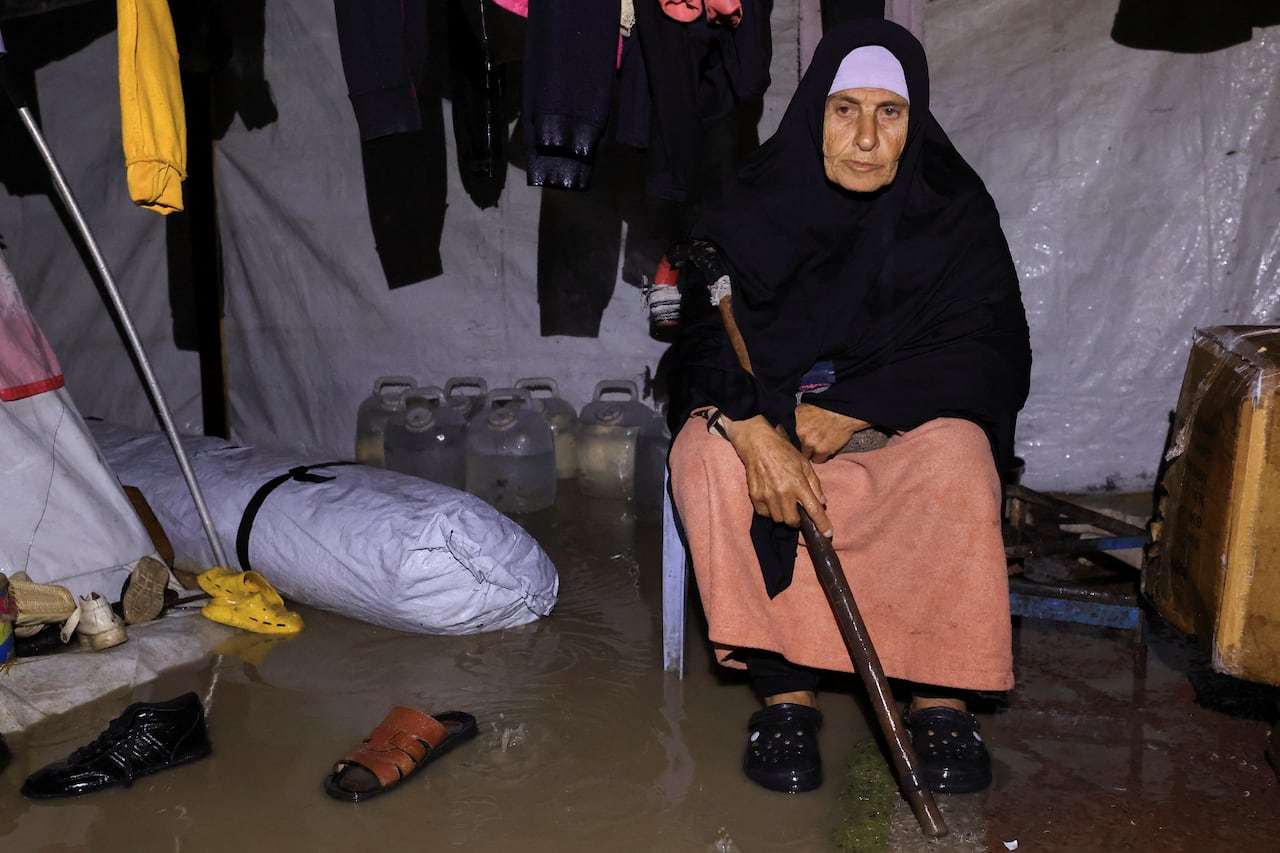 An elderly woman holding a crane sits on a chair in the middle of a flooded tent.