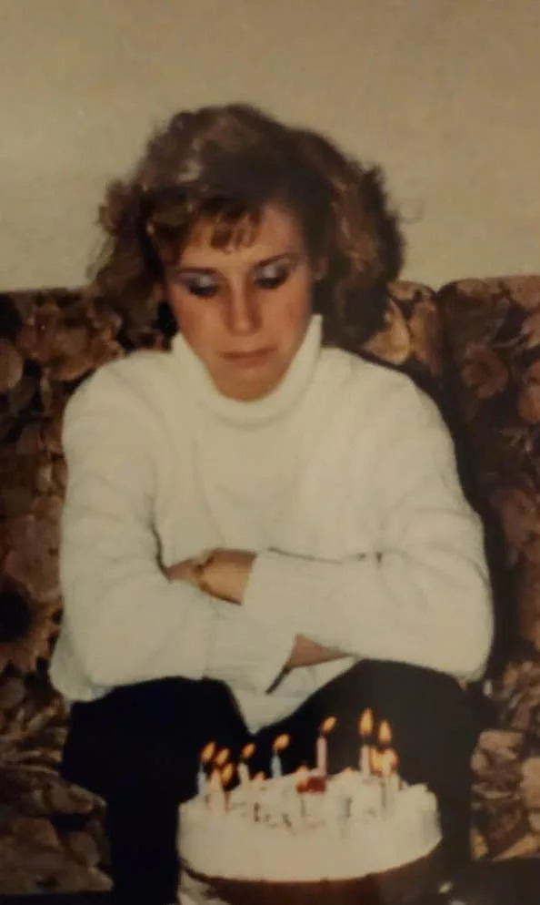 Person in a cozy room seated on a couch, wearing a turtleneck sweater, looking at a birthday cake with lit candles