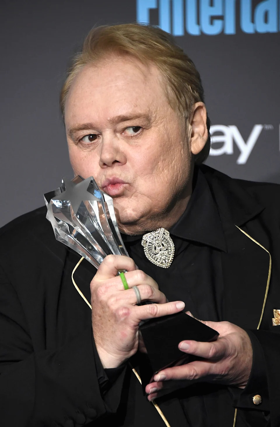 Person in black outfit with embellished collar holding and kissing a star-shaped trophy on stage