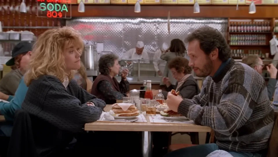Two people sit facing each other in a busy diner, deep in conversation, with food and drinks on the table. Diners are seen in the background