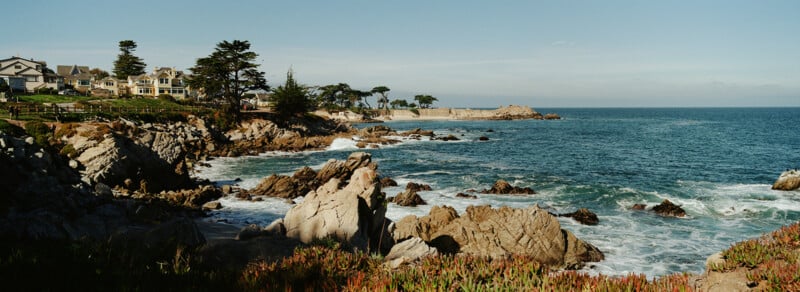 Rocky coastline with waves crashing against the shore, houses and trees on the left, and a clear blue sky above. Lush greenery lines the foreground.