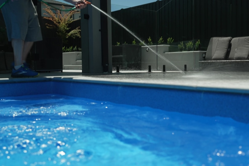 A person holds the orange trigger nozzle of a garden hose and sprays water onto concrete beside the blue water of a pool.