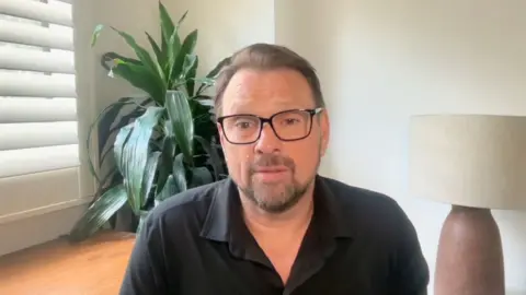A man with a goatee wearing glasses looks into the camera. Behind him is an indoor plant and a lamp