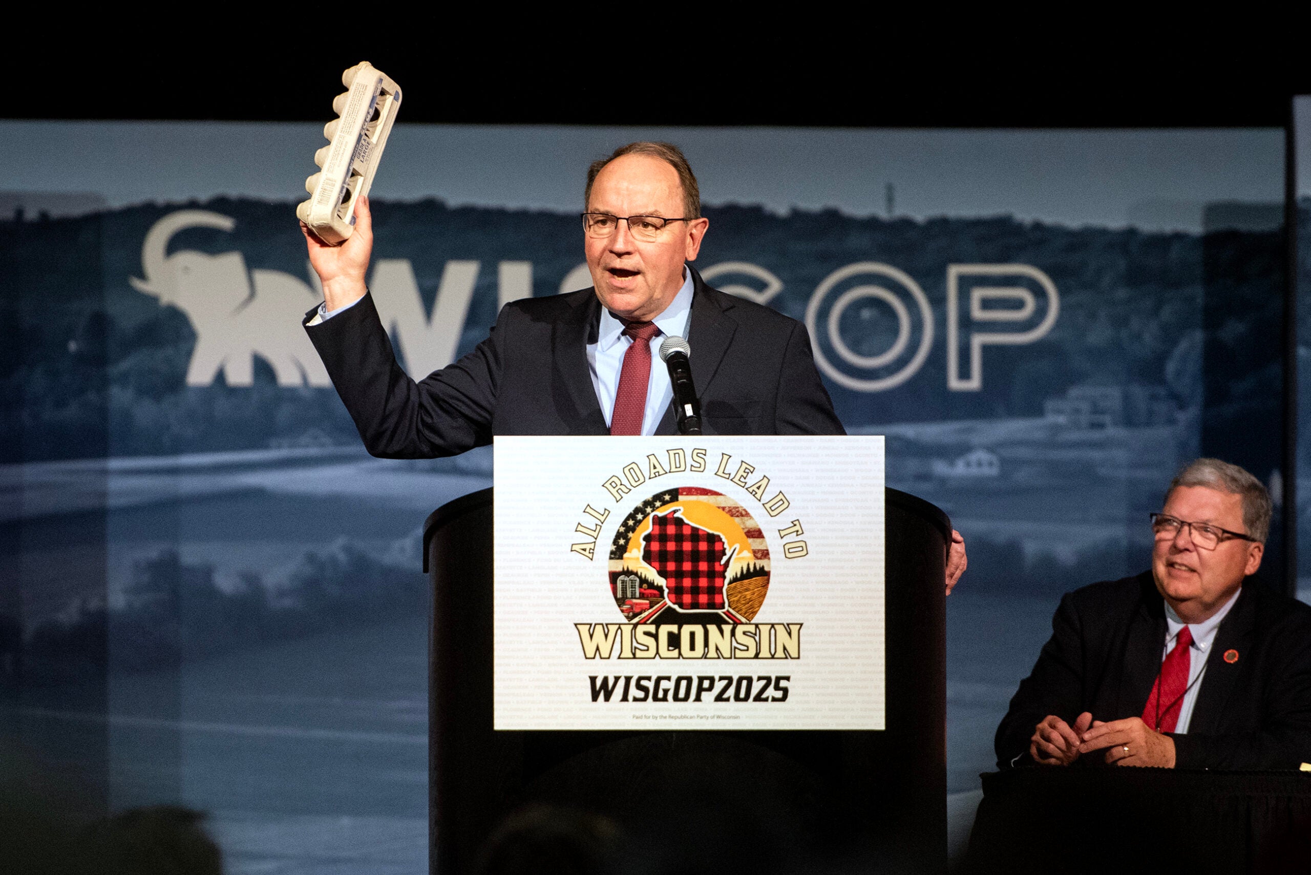 A man in a suit speaks at a podium labeled WISGOP2025, holding an egg carton in one hand, as another man sits beside him on stage.