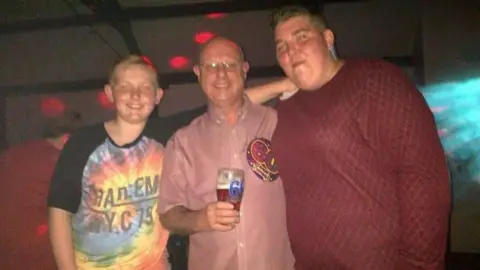 Family handout A boy with short hair stands next to an older man with glasses who is holding a pint of beer and a woman with short hair. All three are smiling.