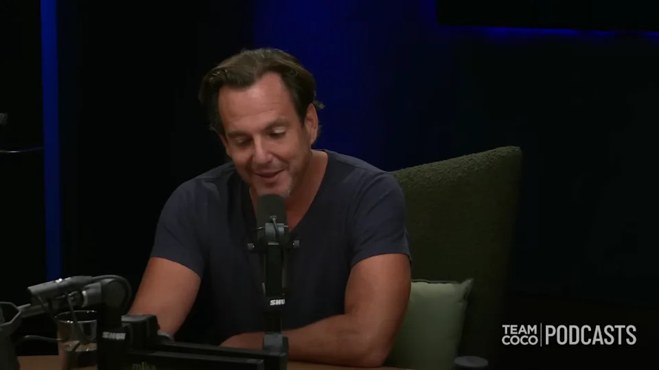 Man speaking into a microphone during a podcast recording, seated at a table with equipment. The setting is dimly lit and casual