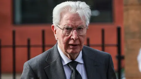SNS Head and shoulders shot of a man wearing a grey suit, white shirt and black tie. He has wavy grey hair and is wearing silver rimmed glasses. Church railings can be seen blurred in the background.  