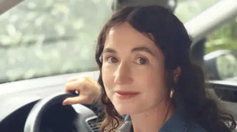 Mary McCarthy A woman with brown wavy hair sat in a car smiling. 