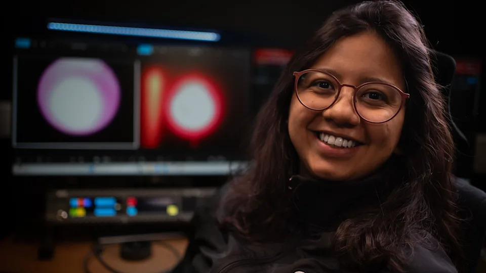 A woman, Veronica Viera, is sitting in front of two large computer monitors in a dark room. The screens show two images of a white and red circular shape - this is plasma glowing in the space furnace. Veronica has long brown hair, she is wearing glasses and a black jacket.