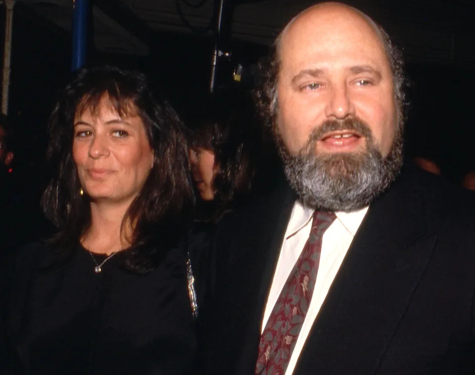 A person with a beard and suit gestures while a woman in elegant attire stands beside him at an event, surrounded by other attendees