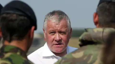 Getty Images Head and shoulders shot of Adam Ingram addressing troops wearing berets and camouflage-style clothing. The soldiers are blurred and have their backs to the camera. Ingram, who has short grey hair combed to the side is wearing an open-necked white shirt and looking to his right.