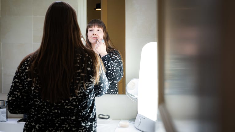 Woman looking into a bathroom mirror and putting on makeup, beside her is a bright light therapy lamp illuminating the room.