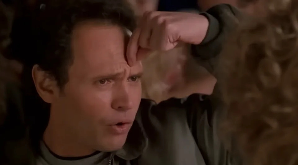 A person with a surprised expression points to their forehead, possibly in disbelief or realization, amid a blurred background of people