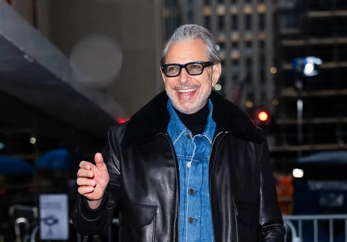 Person wearing denim jacket and pants with a black leather jacket and sunglasses, walking outdoors, smiling and giving a thumbs-up