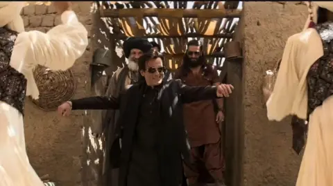 Jio Studios A scene from Dhurandhar movie. Bollywood actor Akshaye Khanna, wearing a black suit and sunglasses, grooves to the music as he enters a brick house, with bamboo ceilings. Two men wearing white robes, sleeveless jackets and turbans dance before him. Bollywood superstar Ranveer Singh, wearing brown kurta and harem pants, and sunglasses, and sporting long hair and beard, along with another actor, sporting a thick beard and black turban, follow Mr Khanna through a corridor leading to the room with the dancers