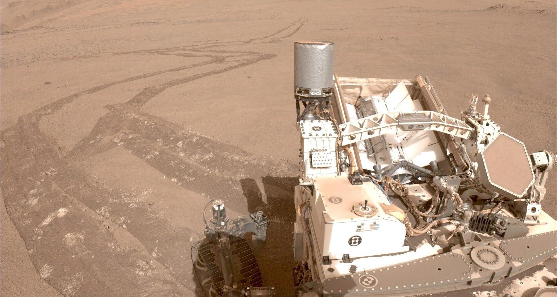 An image taken from one of Perseverance's cameras with the rover in the bottom right of the image and a long windy trail of tire tracks in the reddish brownish dirt of Mars behind it
