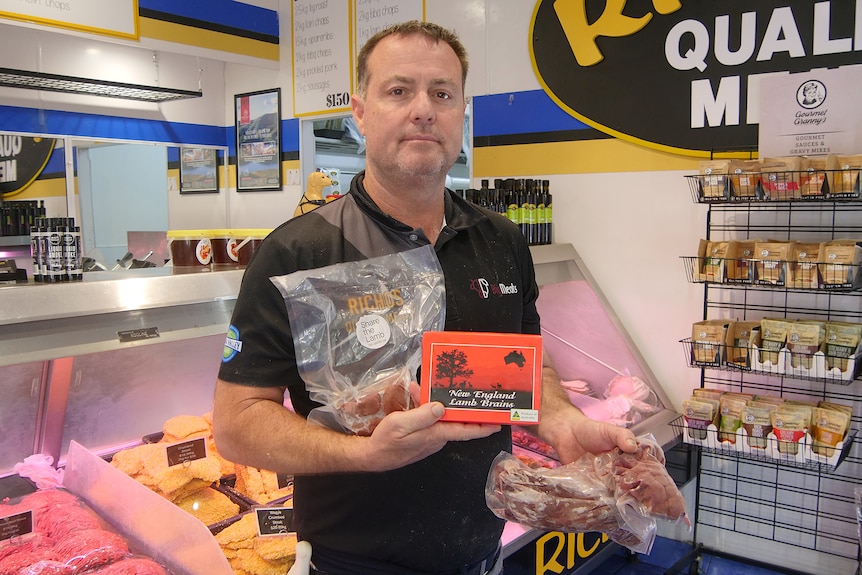 Man dressed in a black collared t-shirt holds up different cuts of meat, infront of a refrigerated display of more meat.