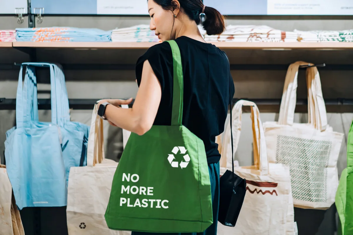 Young Asian woman carrying a green cotton reusable shopping bag with text No More Plastic with a recycling symbol shopping in a store