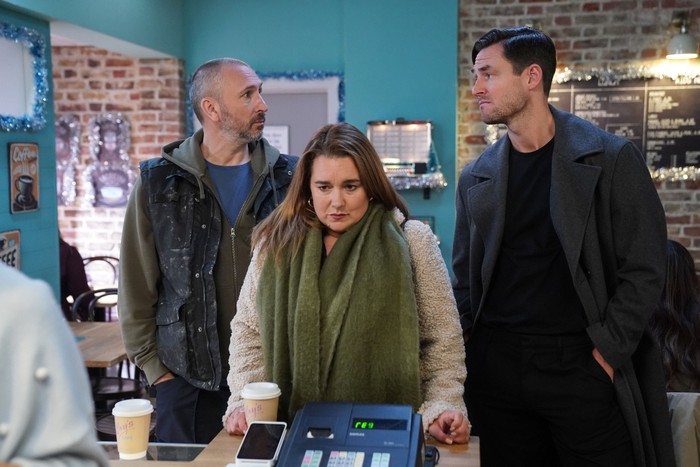 Vicki (wearing a long green scarf) stands awkwardly between Ross and Zack - who are staring at one another - in the caff in EastEnders