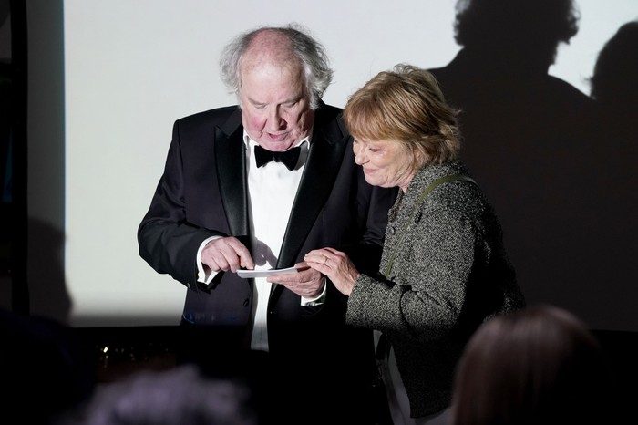 Nigel stands in a bright light wearing a black suit and bow tie beside Julie in EastEnders