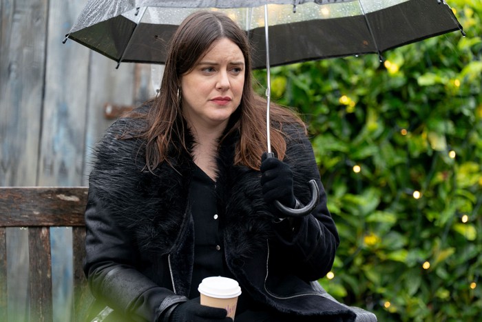 Zoe stands alone under an umbrella holding a coffee cup and wearing a thick black coat in EastEnders
