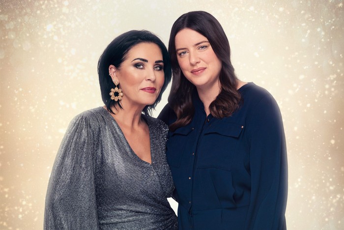Against a golden, glittery background, Kat and Zoe put their arms around each other. Kat is wearing a silvery, glittery dress and Zoe is in a navy blue shirt.
