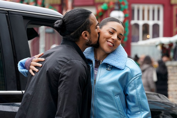 Ravi, wearing a black coat, kisses Priya (who is in a blue coat) on the cheek in EastEnders. They are stood next to a black cab.