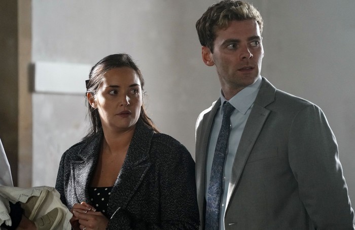Lauren, wearing a grey coat, stands shocked beside Peter, who is in a grey suit, in the church in EastEnders