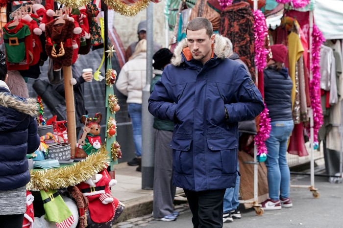 Harry, wearing a blue fur-lined coat, marches through the market in EastEnders