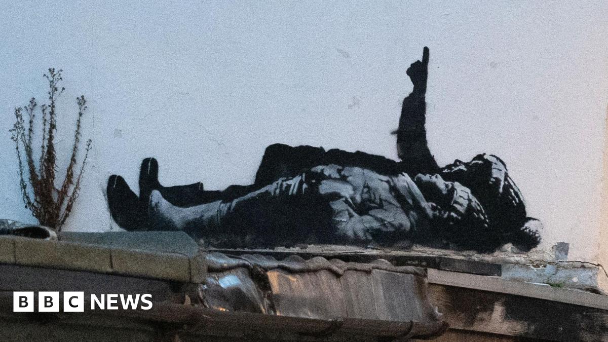 The black and white street art depicts two children lying on their backs and pointing upwards. The child nearest the camera is wearing a wooly hat with hands tucked into their jacket pockets. Behind them a larger person is pointing upwards in a bobbly hat (possibly a Santa type hat) also wearing boots.