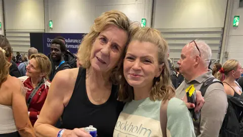 Vicky Edwards Annie Reid stands beside Vicky Edwards and they are smiling at the camera. Mrs Reid holds a can of Red Bull energy drink after her Hyrox competition. Mrs Edwards has short blonde hair and wears a green and white jumper with a rucksack on. People at the Hyrox competition can be seen behind them. 