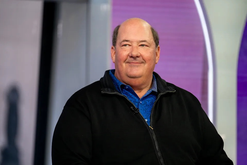 A person wearing a blue shirt and black jacket is seated and smiling during an interview on a talk show set