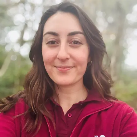 Naomi Bentley of Saving Scotland's Red Squirrels is photgraphed in a bright maroon zip top with the wording 'Saving Scotland's Red Squirrels' on it, smiling at camera, in woods.