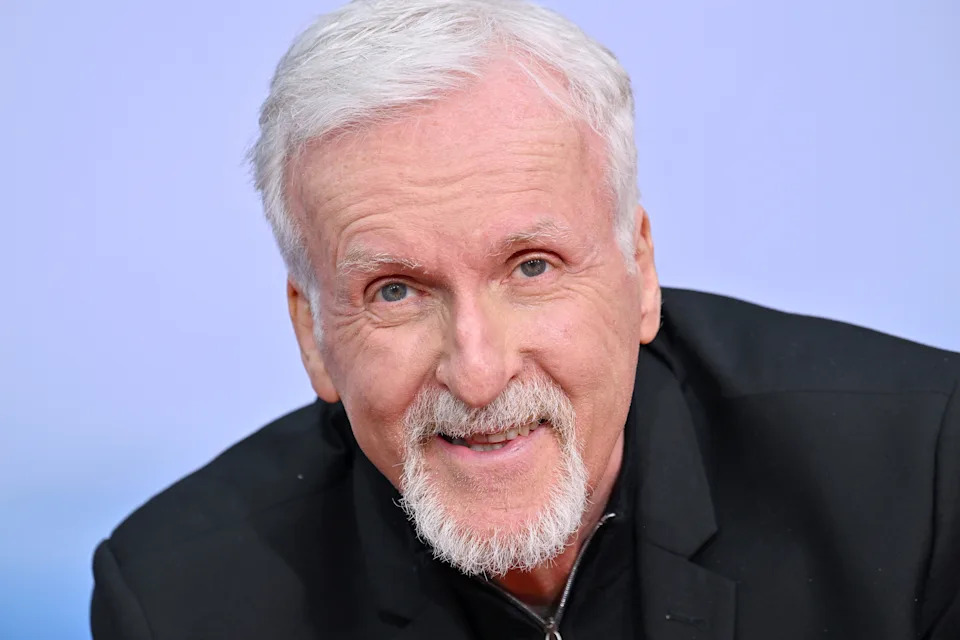An older man with a goatee is smiling at the camera, wearing a black suit jacket over a zip-up top