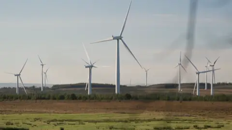 BBC About 10 white wind turbines spinning on a green and brown field site with a grey sky overheard