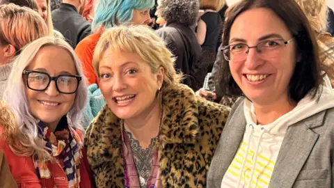 Mitch Schofield Odette Campbell is seen in the middle of a photograph of three women. They are standing together at an event and there is a crowd of people around them.