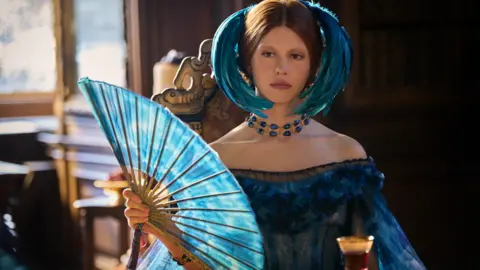 Netflix Actress is Mia Goth, who plays Lady Elizabeth Harlander in the film is sitting at a table wearing an elaborate blue headdress and holding a blue fan. She is wearing a blue dress and there is a glass on a table in front of her