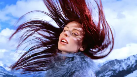 Brit Awards Rose Gray has red hair which she is sprawled in the air as she throws it back over her head. She is wearing a blue fluffy jacket.  