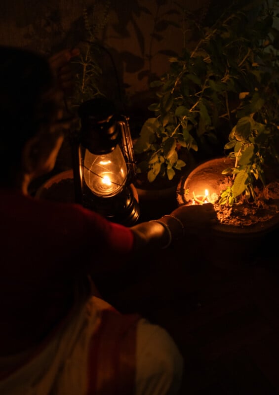 A person in traditional attire lights an oil lamp next to a potted plant, with a lantern also glowing nearby, creating a warm, serene ambiance in a dimly lit setting.
