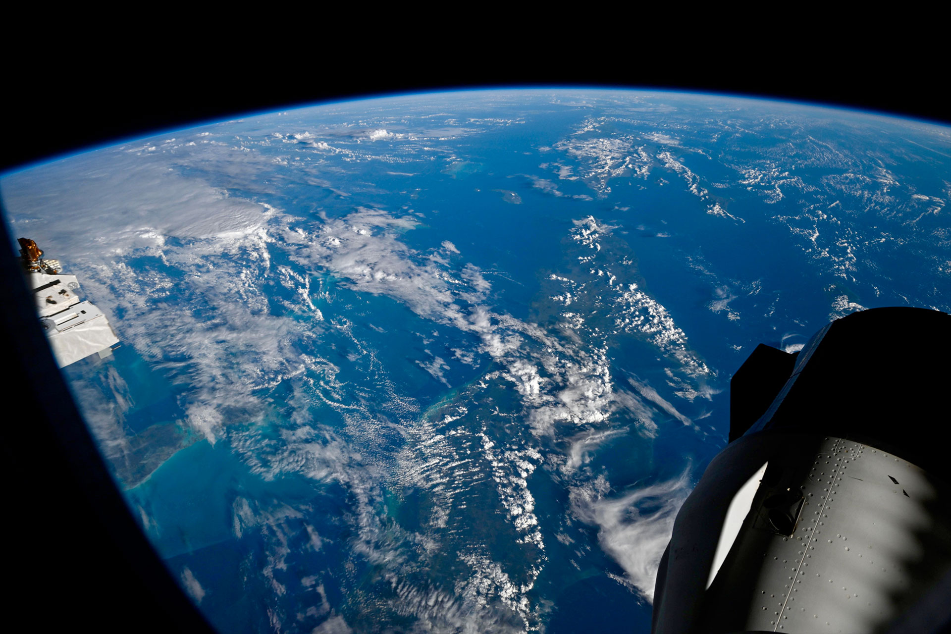 the blue waters and white clouds on Earth extend out to the curved horizon, set against the blackness space and in the foreground, components of a space station