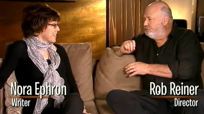 A woman and a man sit on a sofa engaged in conversation. On-screen text identifies them as a writer and a director, respectively