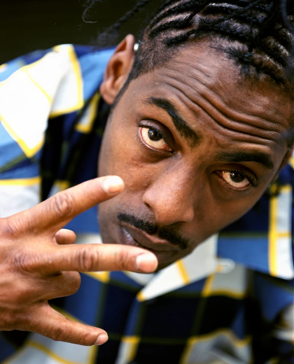 Person with braided hair poses, gesturing with fingers. They're wearing a patterned shirt and looking directly at the camera