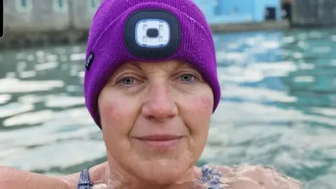 Sam Skyrme-Blackhall Sam is a white woman with blue eyes in a head and shoulders shot of her in the sea. She is wearing a purple woollen-style hat with what looks like a built-in head torch. The background is out of focus but some buildings can be seen next to what looks like a possible harbour or slipway wall.