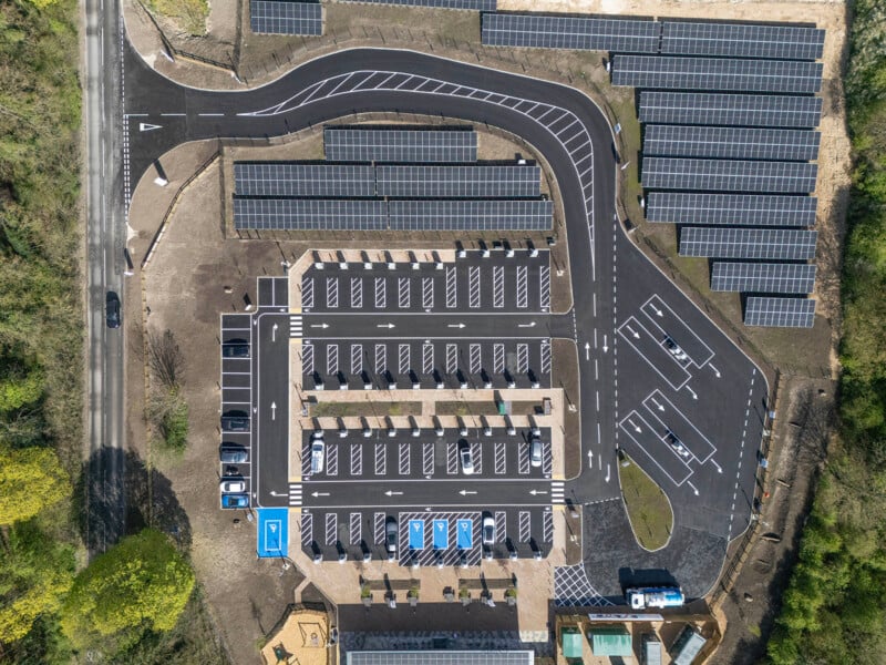 Aerial view of a parking lot with rows of parked cars, solar panels covering many parking spaces, and surrounding green trees. Some empty parking spaces are visible, and there is a lane for cars to enter and exit.