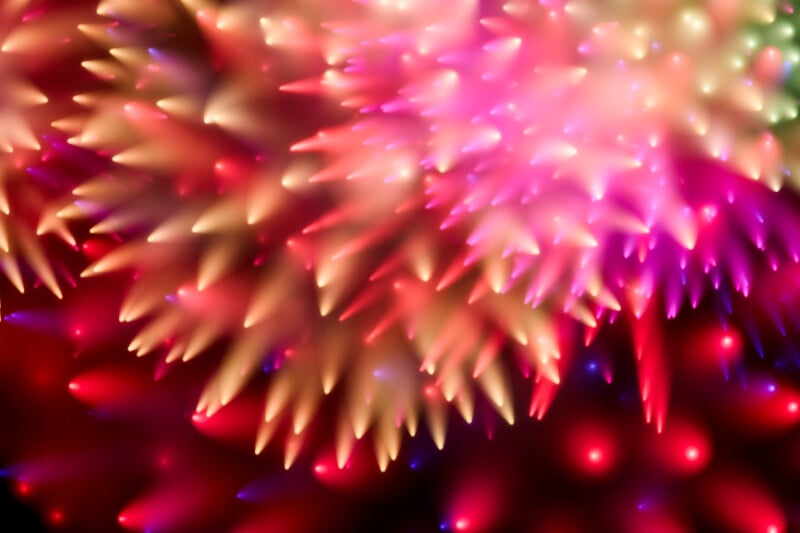 Abstract image featuring vibrant, cone-shaped spikes in pink, red, yellow, and soft green tones, with glowing highlights, creating a dynamic, colorful, and slightly blurred visual effect.