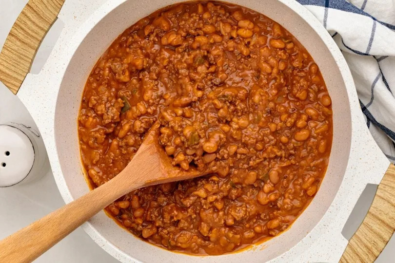 A pot of ground beef and beans being stirred with a wooden spoon, set on a wooden surface next to a blue and white striped cloth and a salt shaker.