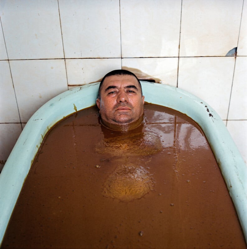 A man lies in an old bathtub filled with thick, brown liquid, partially submerged up to his chest. The background shows stained, white-tiled walls.