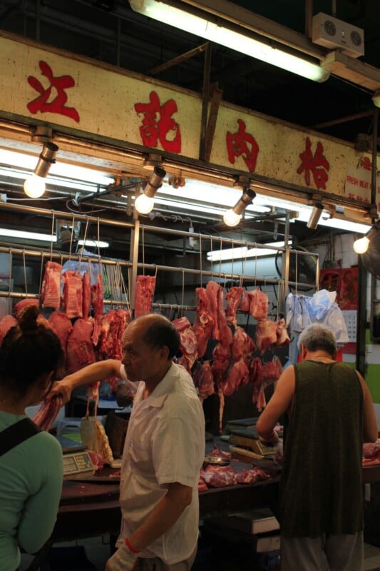 A butcher shop with hanging cuts of raw meat, bright overhead lights, and three people, including a butcher serving customers at the counter beneath a sign with Chinese characters.