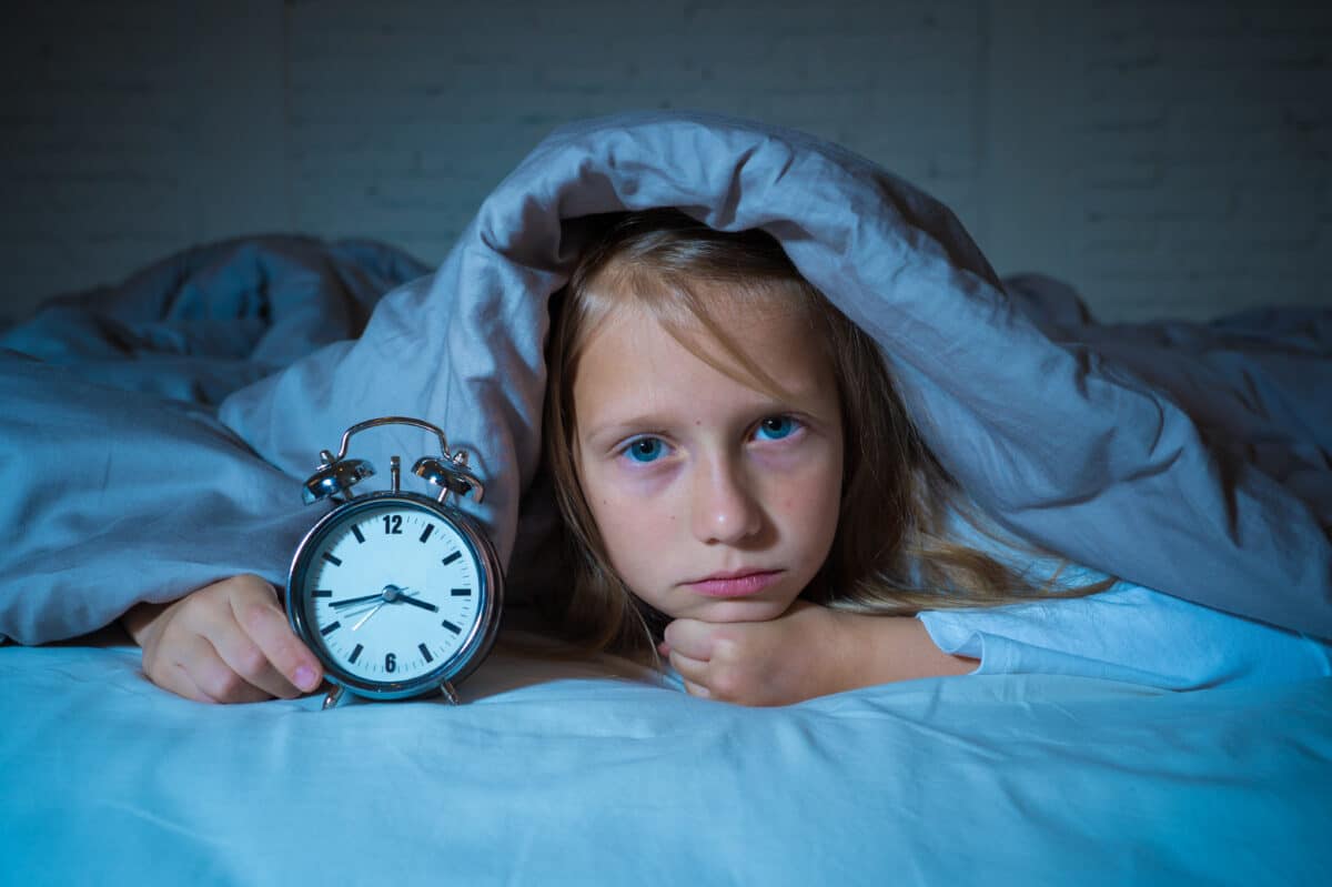 Little girl battling insomnia at night can't sleep and lies awake in bed with a clock
